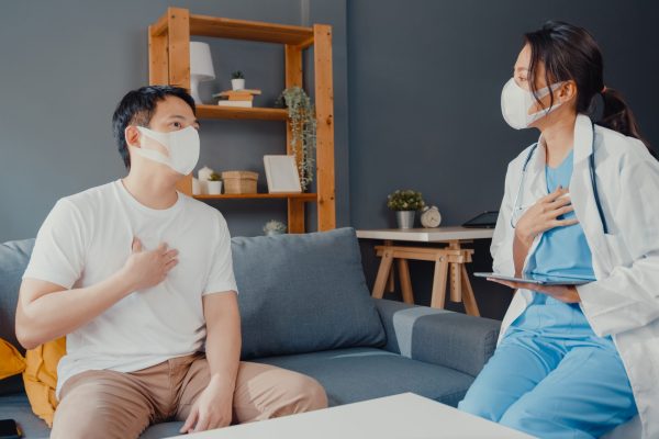 Young Asia female physician doctor wear face mask using digital tablet sharing good health test news with happy male patient sit on couch in house. Medical insurance, Visit patient at home concept.